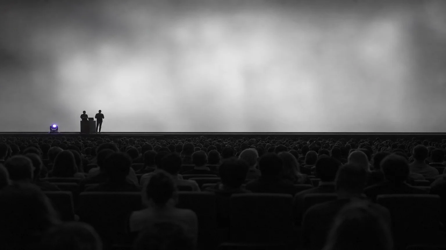 Two engineers presenting from the stage of a large darkened cloud-computing conference hall. Illustrative AI-generated editorial image.